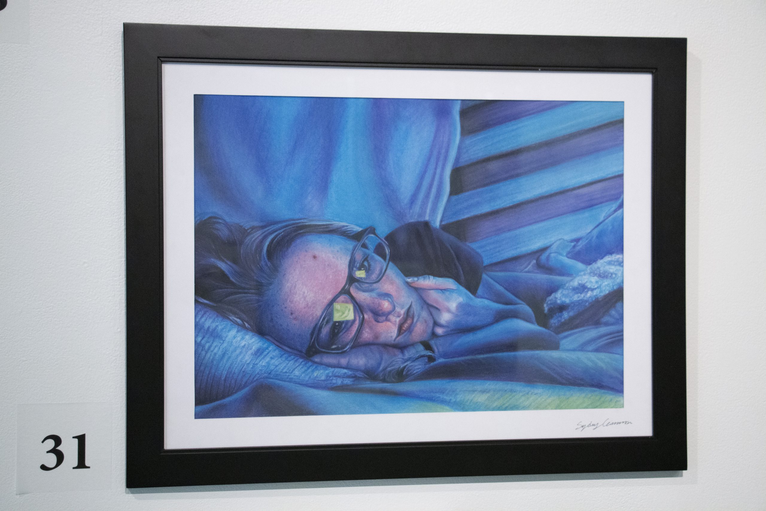 A woman lies on her side in a bed. A reflection in her glasses suggests she is looking at a glowing screen. The entire scene is tinted with a blue glow suggesting that there is no natural light in this photo. Her expression appears lethargic or melancholy.
