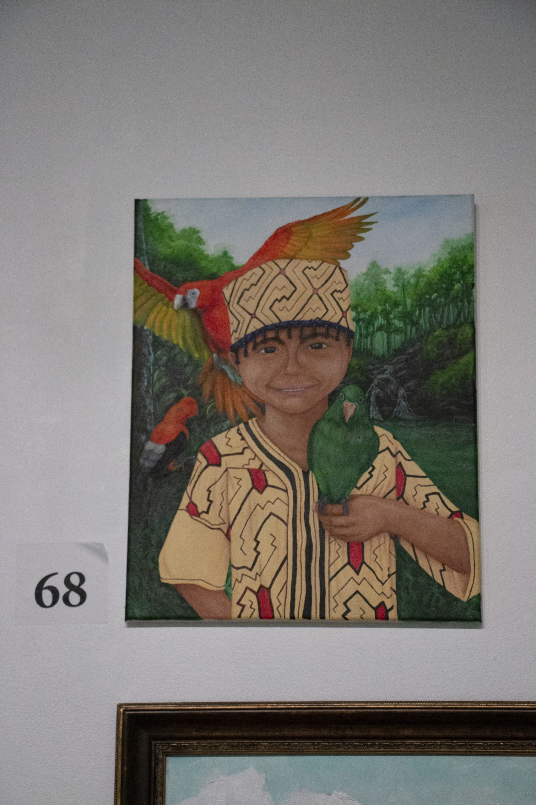 A small child is the central focus. They are wearing a yellow shirt and hat with red and green colorful designs on it. They hold a green parrot in their right hand and a red maccaw spreads its wings behind their right shoulder. The background is lush green forrest.